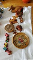 Photo of carved wooden duck, chicken figurines, wooden train set, carved floral box, small wooden container shaped like a house, round painted floral coaster/tray, and small puzzle piece on a white cloth.