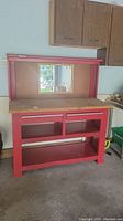 Front view of red metal work bench showing wooden work surface, pegboard back with small mirror, two open storage shelves below work surface, and open bottom shelf.