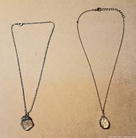 Two necklaces laid flat on a beige surface showing their chains and distinct clear pendants.