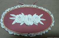 Full top view of the oval crocheted lace wall decoration showing three white crocheted flowers and leaves mounted on red backing within a wooden embroidery hoop frame.