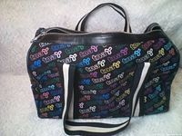 Front side of black fabric TNA Aritzia bag with colorful 'tna' and butterfly logos and black and white striped strap shown lying flat.