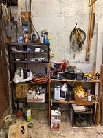 Two shelves against a concrete block wall, containing automotive chemicals, electrical cords, tools and various containers of oils and coatings.
