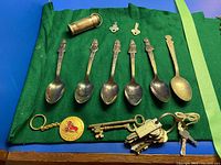 Photo of 6 silver plate souvenir spoons, one gold-tone round keychain with red rose emblem, brass-colored dime keeper capsule, and several keys on two keyrings plus a tiny skeleton key, all displayed on green fabric.