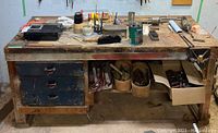 Wide view of the metal workbench with tools, including drawers, clamps on top, containers with small parts, and various tools scattered.