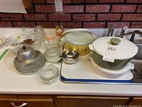 Full lot of kitchen items shown on counter and in sink including Corning Ware, Pyrex casseroles, various glass bowls, metal butter dish, and salad servers.