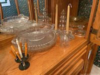 Crystal and glass bowls, plates, candle holders with taper candles, pinwheel crystal bell, and cutlery set in stand on wooden shelf.