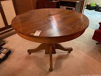 Side angle showing round wood tabletop with visible grain and finish wear, plus pedestal base with four legs ending in casters.