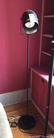 Full view of the black metal floor lamp with round base and adjustable flood light style head, standing against a pink wall.