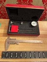 Set inside a foam-padded box including dial indicator, clamps, pressure gauge, rods, and metal caliper on table with ruler for scale.