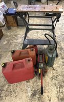View of the Workmate deluxe portable work center, three red jerry cans, red trolley jack, and compressed air sprayer on garage floor.
