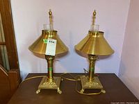 Two matching brass-tone adjustable metal lamps with wide glass shades, sitting side by side on a wooden surface, showing square claw-foot bases and electric cords.