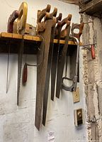 A row of approximately seven hand saws hanging on a wooden rack on wall, including coarse and fine toothed blades with wooden handles.