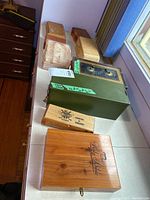 Nine storage boxes including two green metal locked boxes, multiple wooden cigar boxes, a wooden Bible holder, and a vintage plastic jewelry box, displayed on a table.