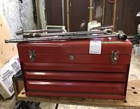 Red metal toolbox with lock and three drawers with several lug wrenches and socket extensions lying on top of it.