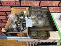 Overall view of both vintage metal grinders in a cardboard box next to three metal baking pans including a muffin tin and rectangular pans.