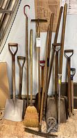 Full view of assortment of yard tools leaning against wall including shovels, broom, claw tool, hoe, and replacement handles.