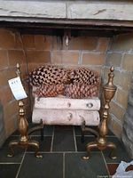Pair of brass andirons positioned in a fireplace, showing their height, finish, and design including finial tops and three-legged bases.