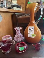 Photo showing five art glass items including an amber pitcher, red and white hobnail vase, red ashtray, hobnail dish, and bird figurine on wooden surface.
