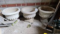 Three large cement pots with visible chipping and cracks displayed against a brick wall on a concrete floor.