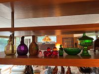Shelf view of assorted colored glass decanters, bell, faux fruit, bowl, and pedestal dish with lid.