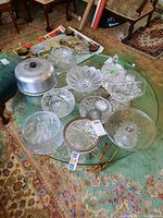 Overview of the lot showing multiple crystal bowls and canisters with pinwheel cut patterns on a glass table, along with the silver-toned cake stand with dome lid.