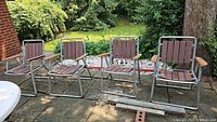 Four lawn chairs placed outdoors on a stone patio. All have wood slatted back and seat panels, silver metal frames, and wooden armrests.