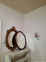Photo showing two framed decorative wall mirrors; one with ornate gold frame and one with round wooden frame, both resting on a shelf.