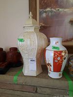 Tall cream-colored ceramic canister with lid and raised relief designs alongside a white vase with orange-red and floral patterns, both on a shelf.