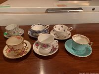 Six teacups with matching saucers displayed on wooden surface. Various floral patterns and gold trim visible. Some visible chips.