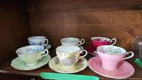 Wide view showing six assorted teacup and saucer sets of various colors and floral/scenic designs on wooden shelf.
