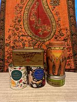 Five empty decorative tin boxes arranged with a colorful tapestry background. Shows various designs including elephant, butterflies, botanical and vintage script.