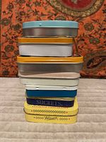 Stack of seven vintage tins with visible brand labels including Sucrets and Whitmans on the bottom tins. The tins are stacked with a patterned textile background and carpet floor allowing clear view of size and color.