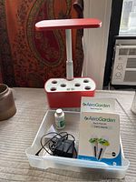 Full setup of AeroGarden 6 pod hydroponic growing machine with red base, white pod holes, and adjustable light panel. Clear plastic tray in front contains power adapter, liquid plant food bottle, and AeroGarden Seed Pod Kits.
