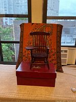 Colonial style wooden doll chair with arm desk on a red box and orange patterned backdrop viewed from front angle