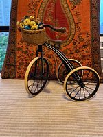 Photo of black metal Samantha doll tricycle with three white-rimmed wheels, brown seat and handlebars, and woven basket filled with yellow and blue artificial flowers, set against a patterned fabric background.