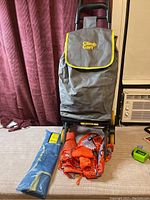 Front view of Climb Cart with gray main bag attached, two orange nylon laundry bags and one blue foldable bag lying on carpet in front.