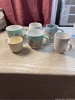 All five coffee mugs arranged on a textured fabric surface with a burgundy curtain background, showing assortment and scale.