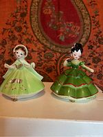 Two porcelain figurines standing side by side, green dresses, one figurine missing shamrock accessory.