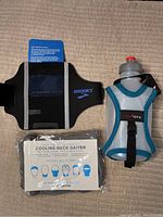 Top-view photo showing Brooks black armband with reflective strips and logo, Ultraspire handheld water bottle in mesh pouch with zipper and strap, and packaged cooling neck gaiter with illustrations.