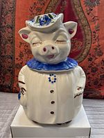 Front view of Shawnee Pottery Winnie Pig cookie jar with blue collar and white coat, smiling pig face with closed eyes, blue and green floral hat.