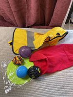 Photo of two small dog coats (yellow raincoat and red hoodie) with four dog toys (rope ball, goofy sounds ball, purple ball for treats, blue wobble ball) and a small green frisbee on a bed.