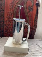 Stainless steel coffee press standing on a white box, showing full height with plunger extended and large handle.