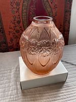 Front angled view of pink frosted molded glass vase with raised floral and leaf design. Vase sits on a white box with a patterned textile backdrop.