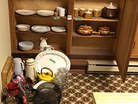 Wide view of wooden cabinet with assorted dishes, soup tureens, kettles, coffee pots, mugs, and glassware arranged on shelves and floor.