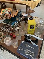 Overview of various bar accessories, including wine openers, corkscrews, decanters, bottle stoppers, and a glass with measurement markers, all laid out on a table.