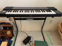 Front view of Lowrey V-100 keyboard on stand showing keyboard, foot pedal, and power cord on carpeted floor with surrounding items visible.