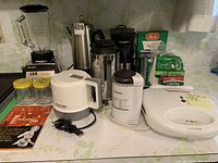 Full spread of kitchen appliances on counter showing blender, coffee maker, grinder, percolator, kettle, sandwich maker, coffee thermos, and coffee filters