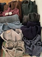 Thirteen men's sweaters in varied colors and textures laid out together showing knits, buttons, and collar styles.