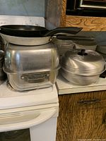 Kitchen countertop with aluminum roaster, rectangular double boiler, and egg poacher with lid. Frying pans stacked on top of the double boiler.