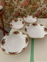 Four Royal Albert Old Country Roses fine china bowls set on a white table with floral wallpaper background, showing the decorative rose patterns and gold edges.